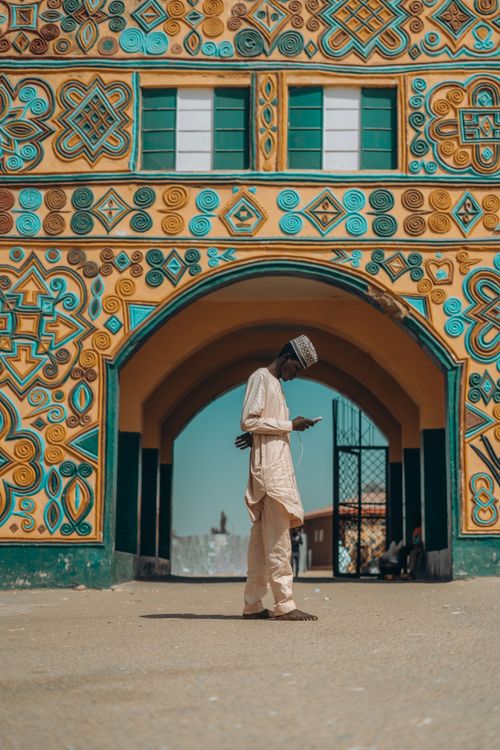 Free Man in traditional attire using a smartphone in front of colorful architecture. Stock Photo