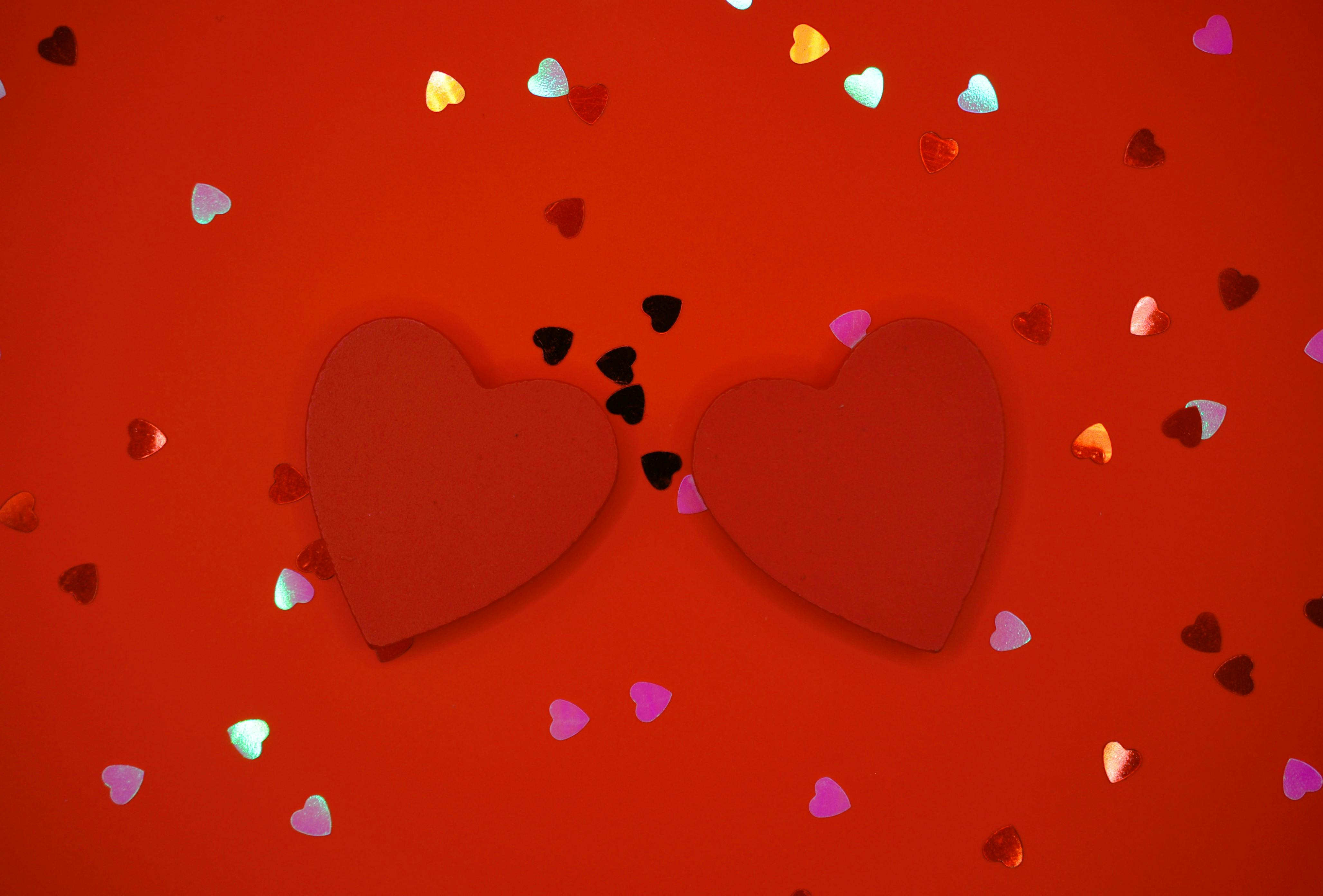 Gratis Due cuori rossi circondati da coriandoli colorati su uno sfondo rosso, simbolo dell'amore. Foto a disposizione