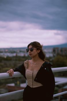 Woman wearing sunglasses and black jacket on a rooftop at dusk with a cityscape backdrop.