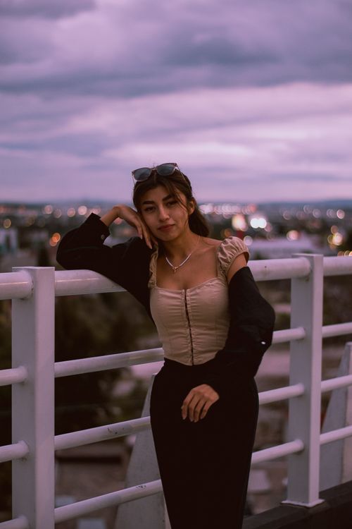 Free Young woman in fashionable attire posing at sunset on a rooftop with a city skyline backdrop. Stock Photo