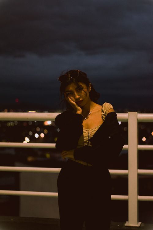 Free Young woman leaning on a railing, city lights twinkling in the background at night. Stock Photo