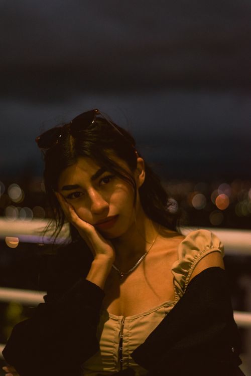 Free A contemplative portrait of a woman against a dreamy city night skyline with warm tones. Stock Photo
