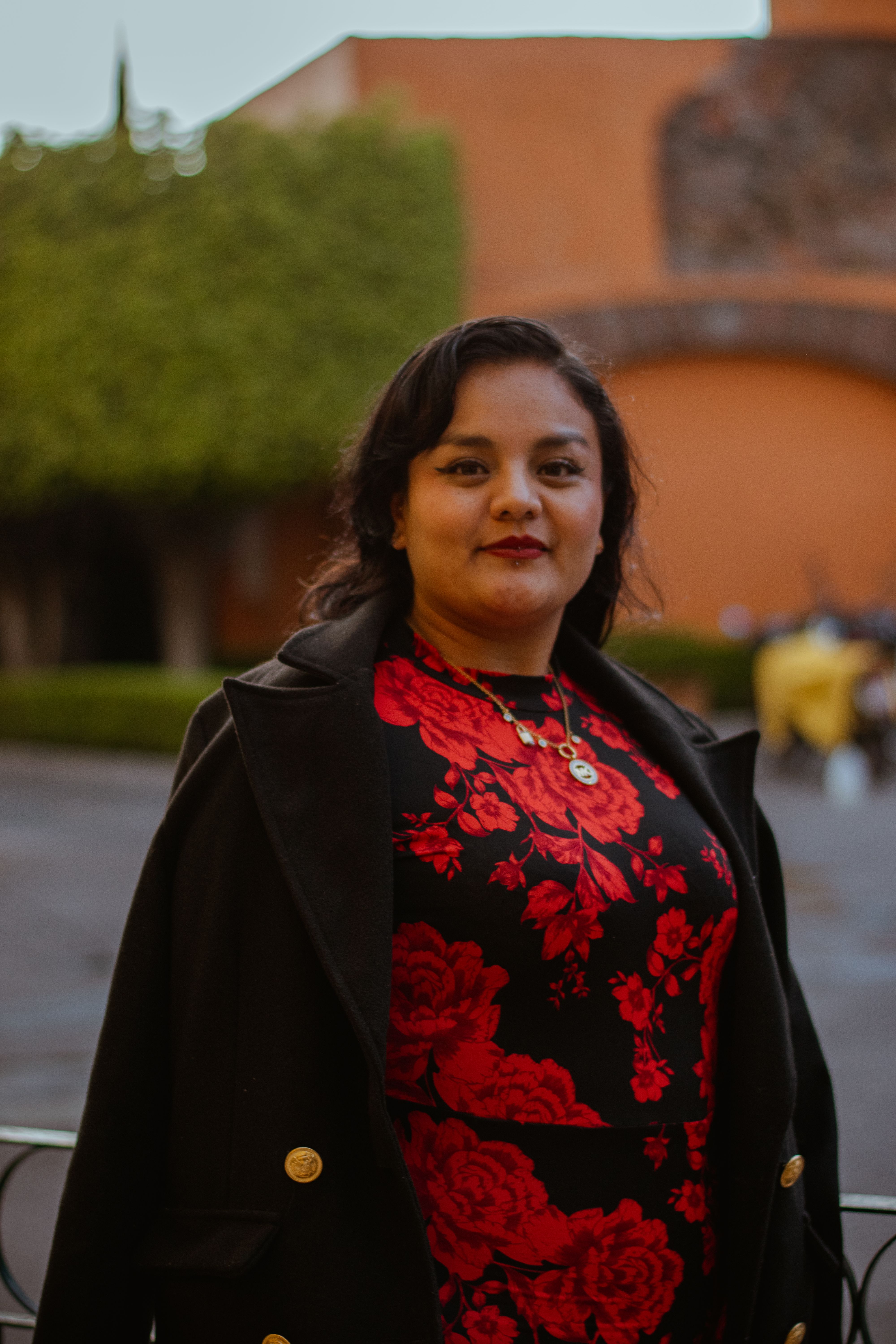 Confident woman in a red floral dress poses stylishly with an elegant outdoor architectural background.