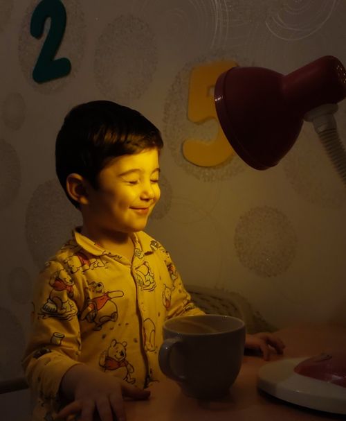 Free A child in pajamas smiles as a lamp casts a warm glow on them, creating a cozy ambiance. Stock Photo