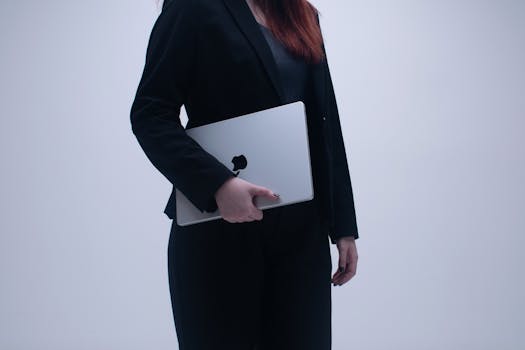 Businesswoman in formal attire holding a laptop against a plain background.