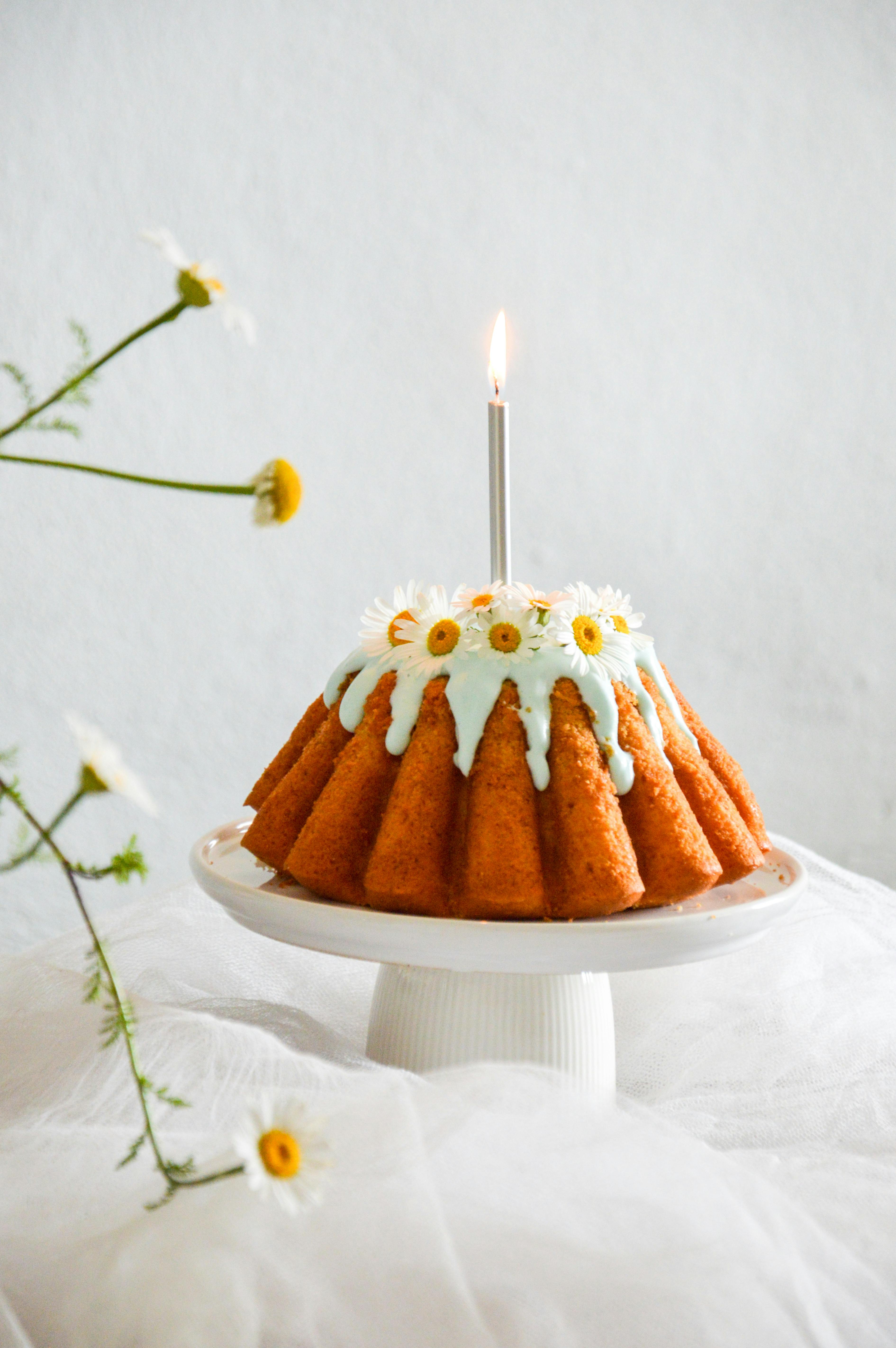 Bundt cake adorned with icing and daisies, featuring a lit candle.