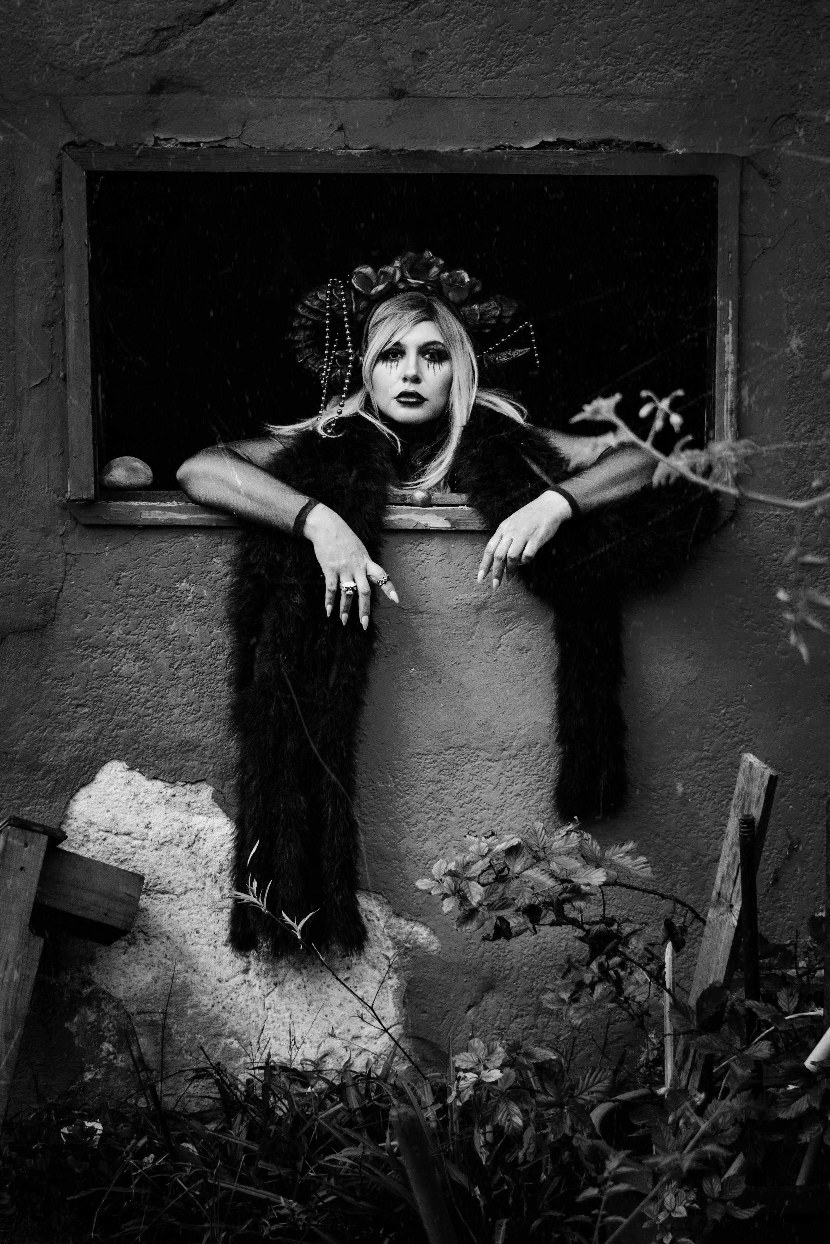 Black and white portrait of a woman in gothic makeup, framed in a window, exuding a dark and moody ambiance.