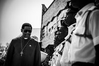 A religious leader walks alongside uniformed individuals outdoors in a monochrome photograph.