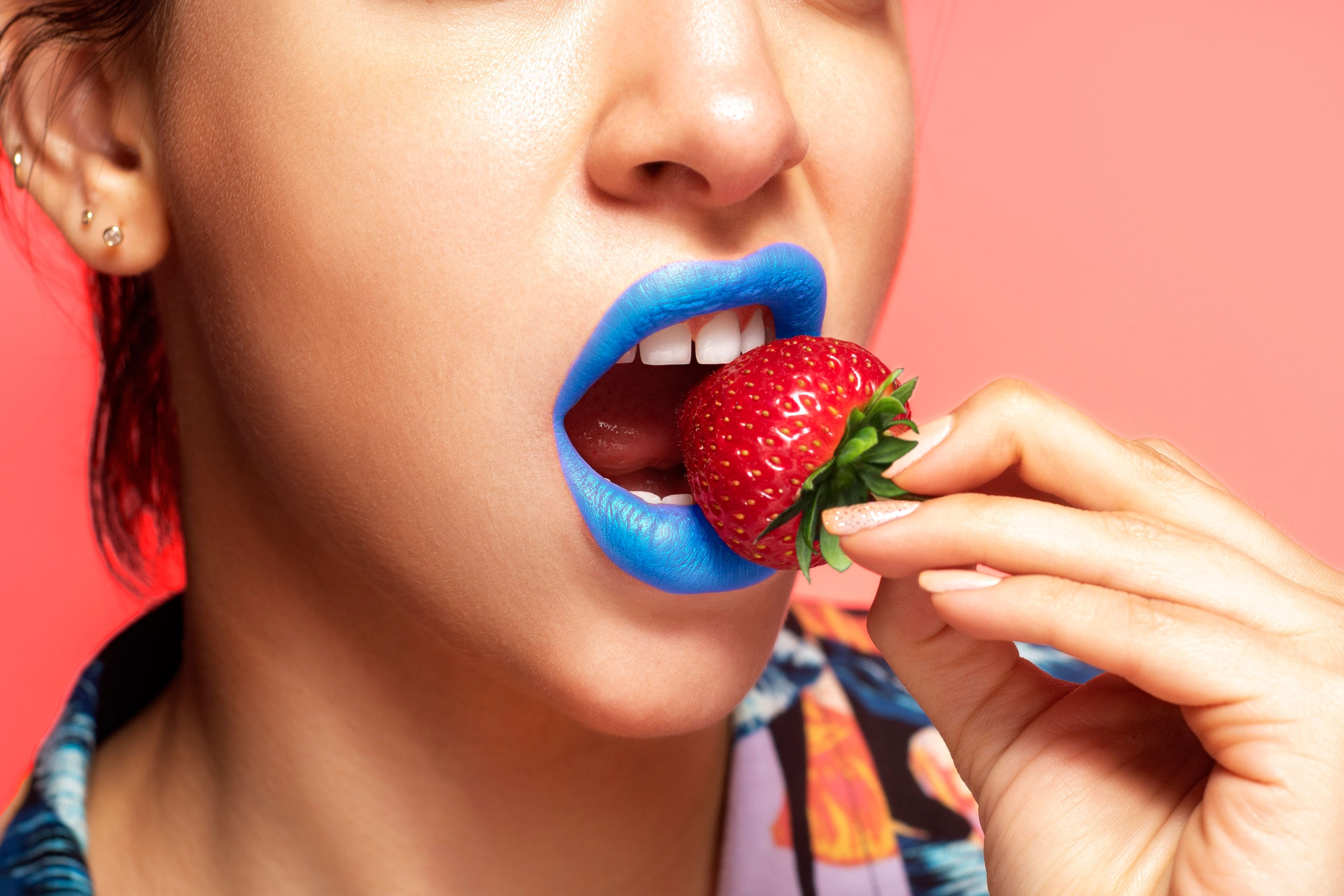 Close-up portrait of a woman with blue lipstick holding a fresh strawberry.