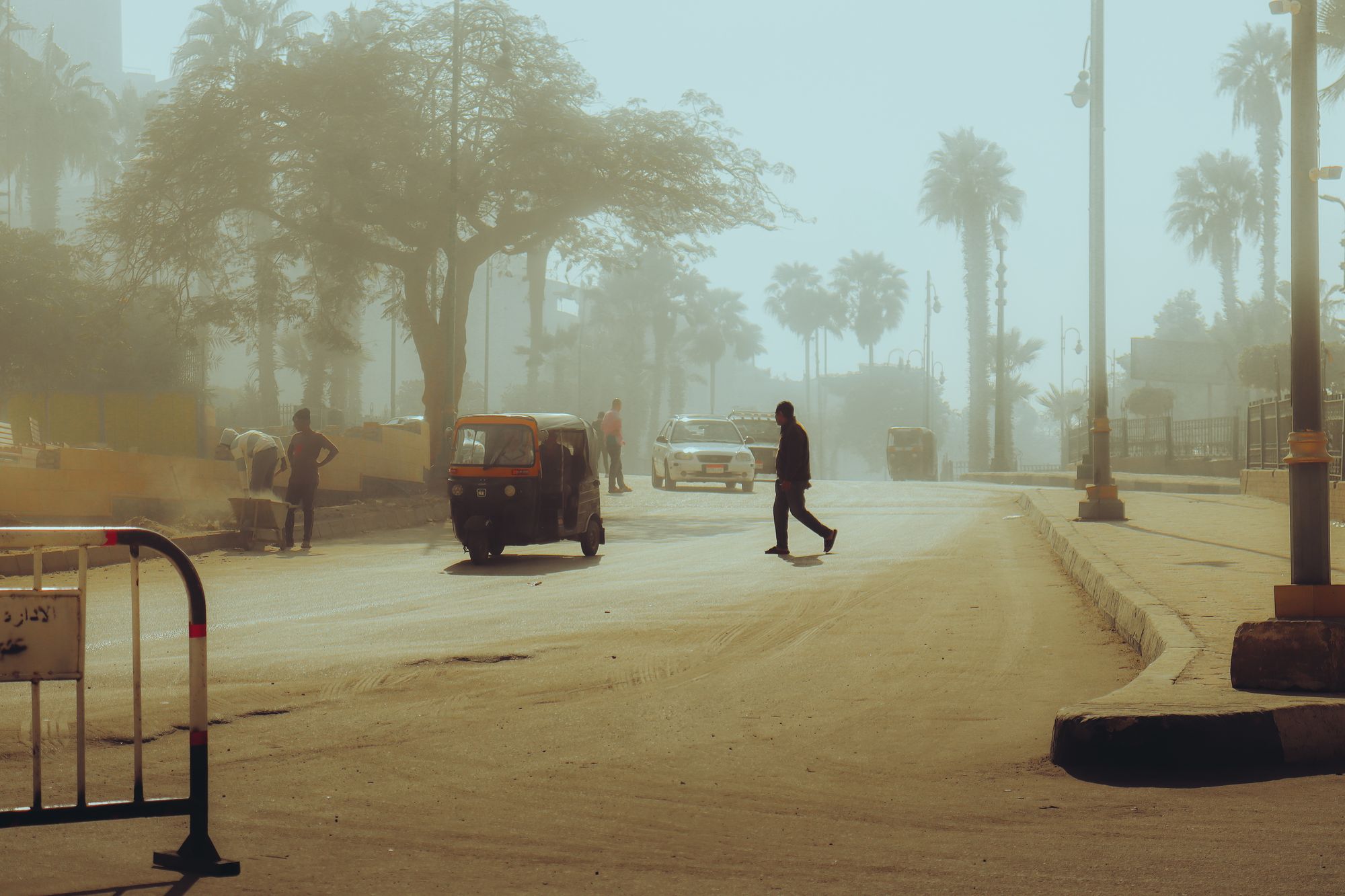 A hazy street scene with a tuktuk, trees, and people crossing.
