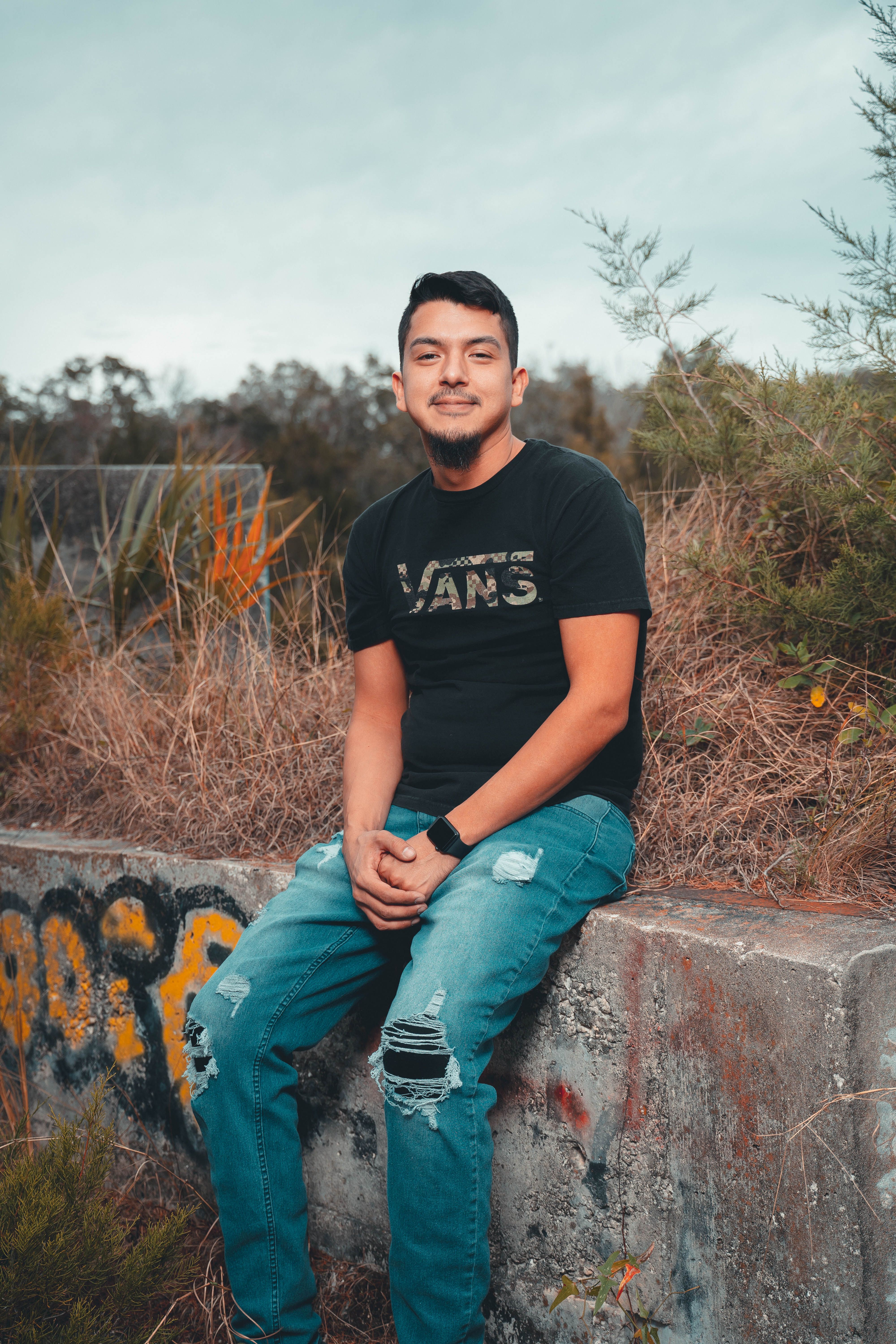 Man wearing black t-shirt and jeans sitting on graffiti-covered concrete outdoors.