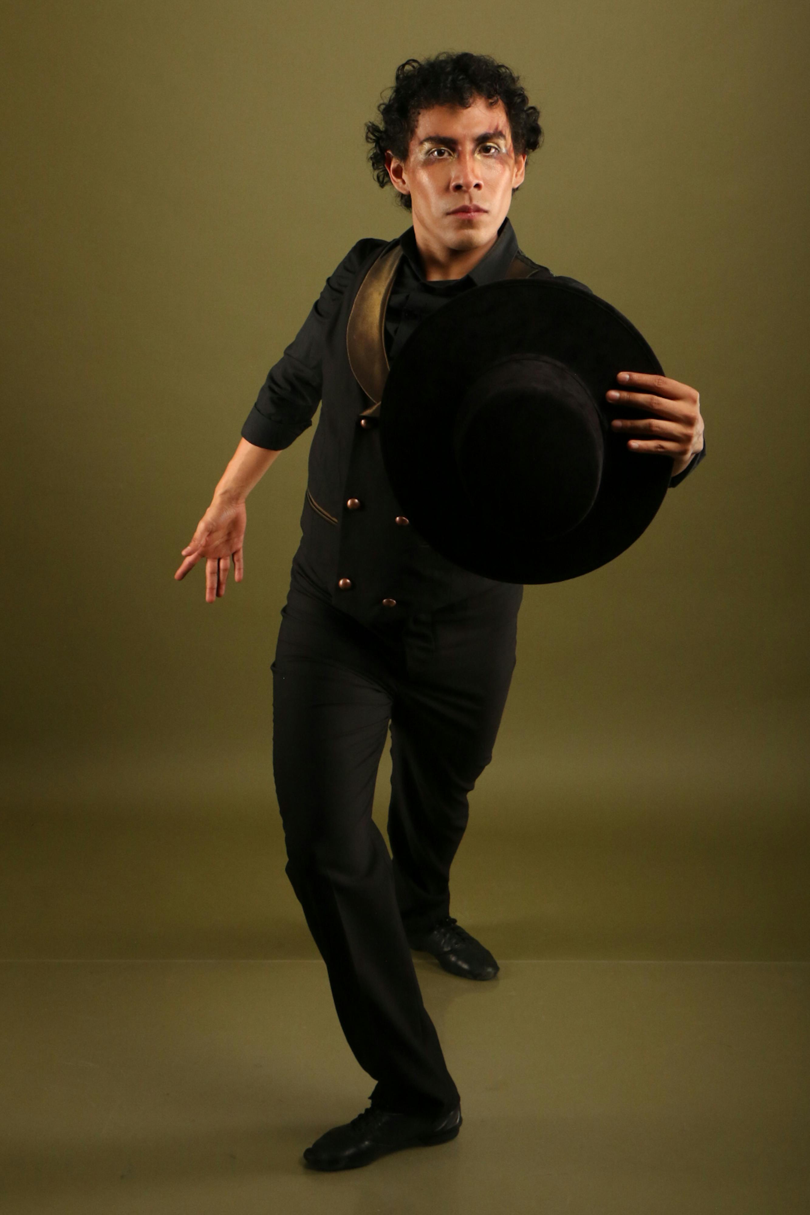 Male dancer in stylish attire performing with black hat against olive background.