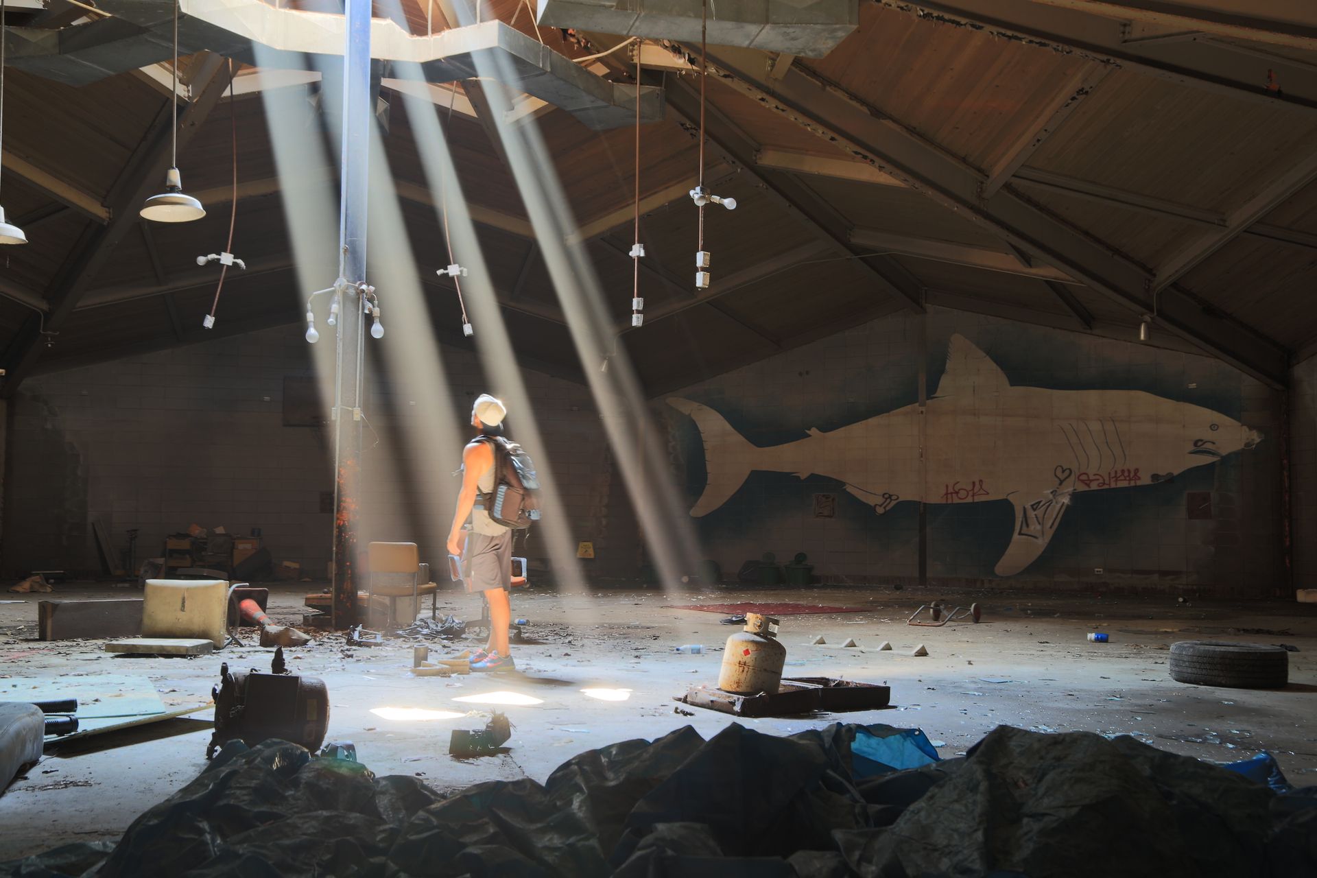 An urban explorer stands in sunbeams inside an abandoned warehouse with a graffiti shark mural.