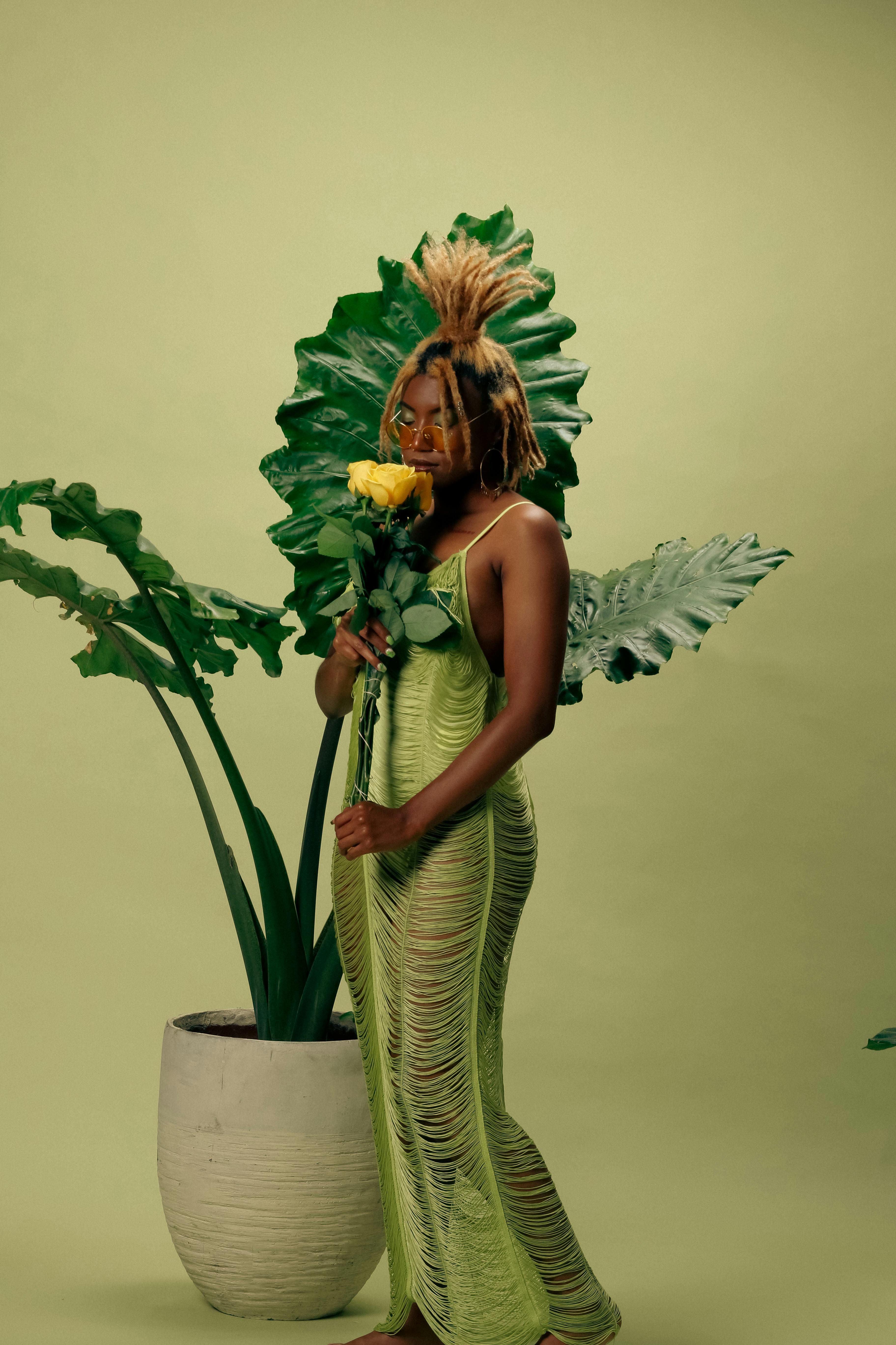 Stylish woman in green dress holding yellow rose against leafy backdrop.