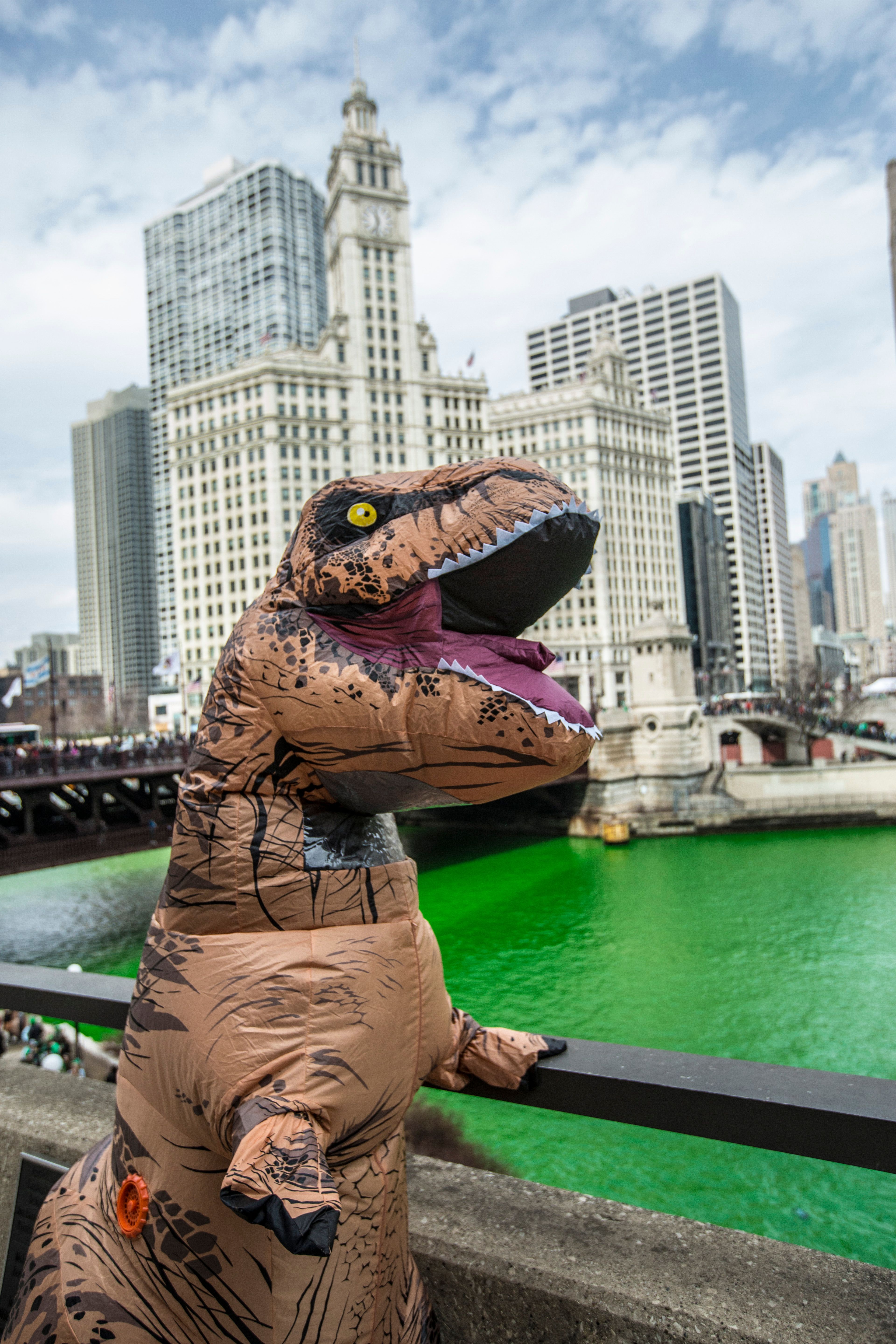 A person in a dinosaur costume poses by the green Chicago River with skyscrapers in the background.