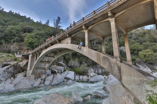 Free A person sitting on a bridge over a rushing river surrounded by lush forest. Stock Photo