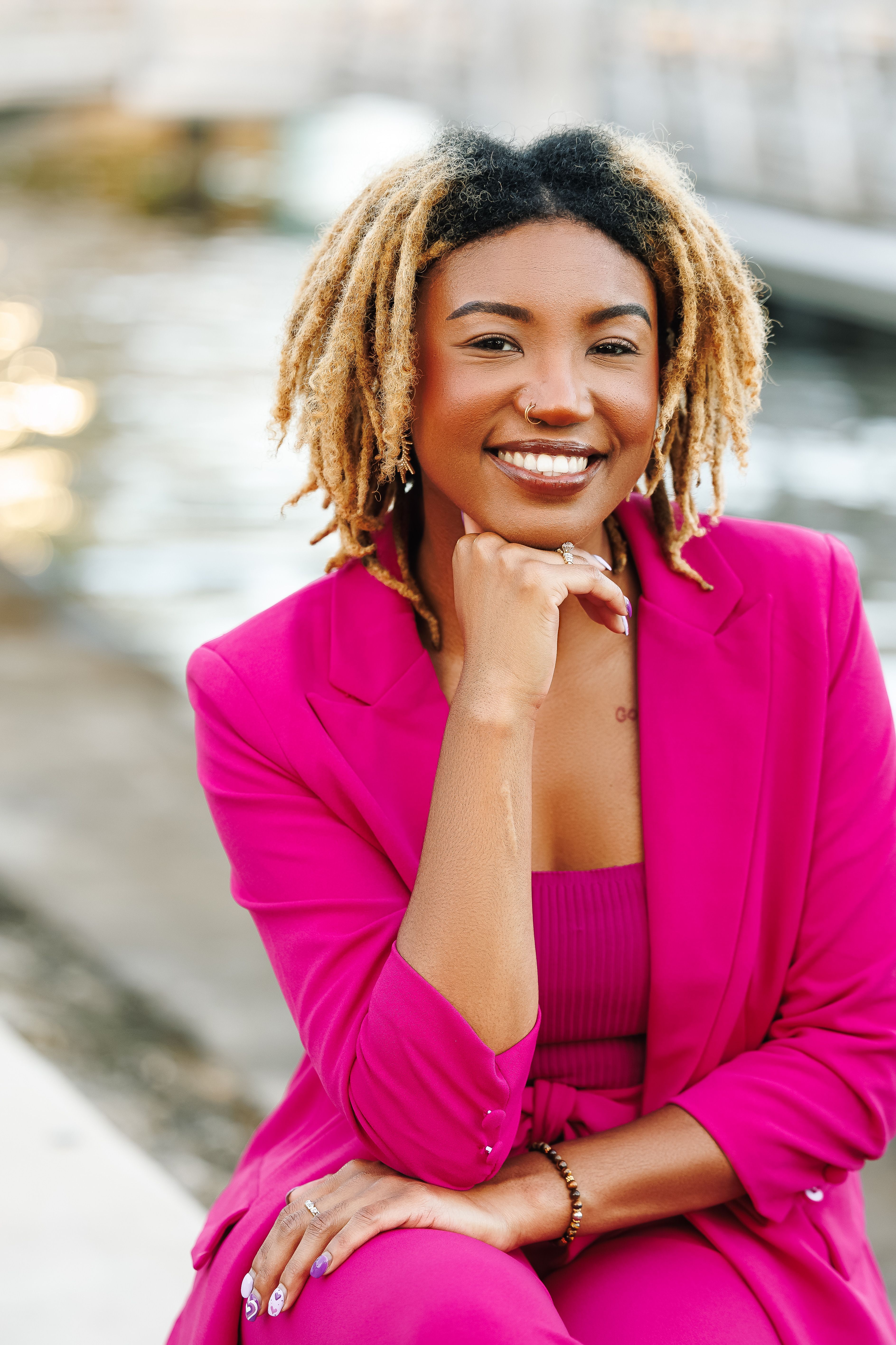 Confident woman in a pink suit smiling by the water, exuding warmth and positivity.