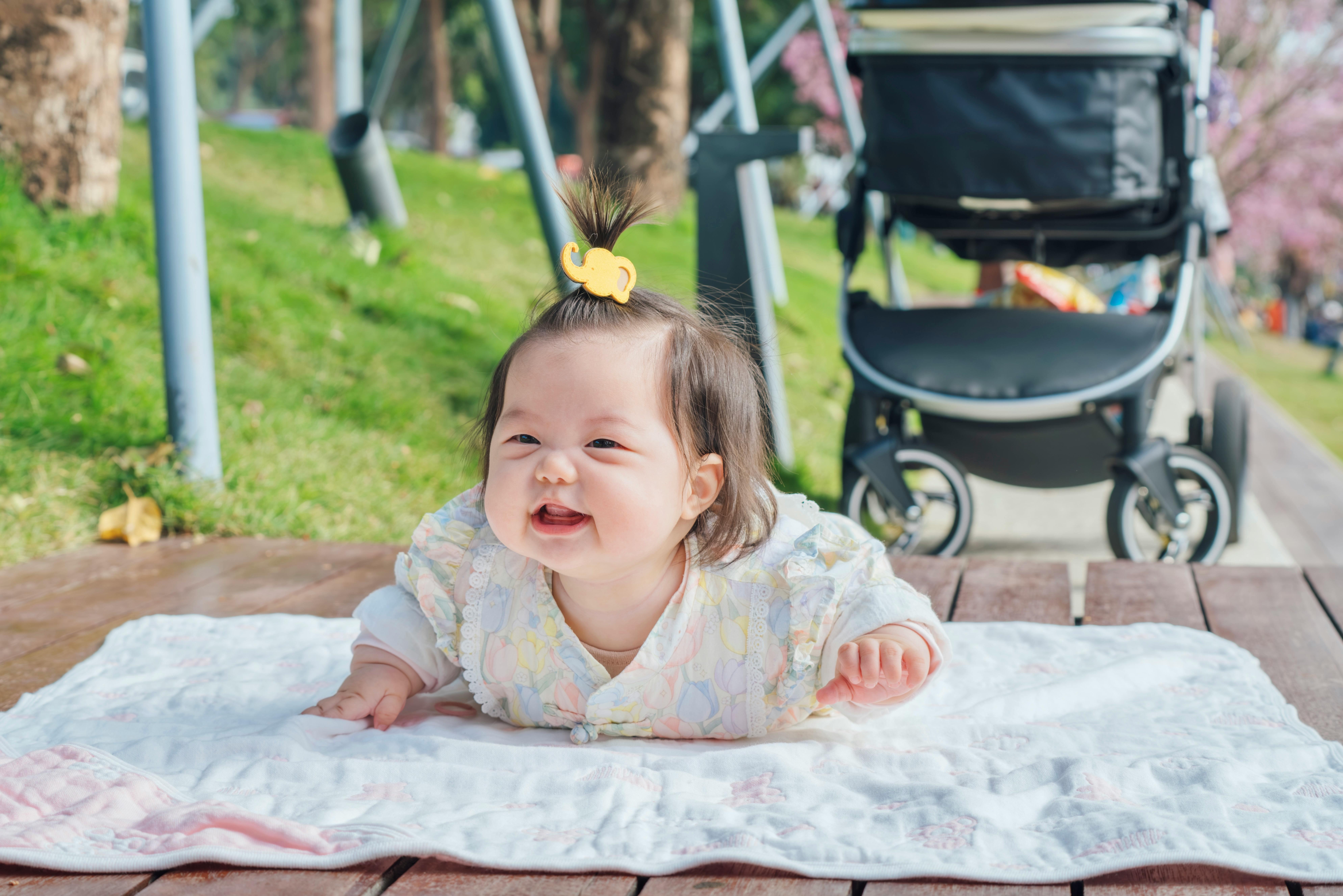 Smiling Asian baby lying on a blanket outdoors beside a stroller on a sunny day.