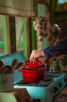 A hand lifts the lid of a red pot on a vintage stove, evoking a nostalgic cooking atmosphere.