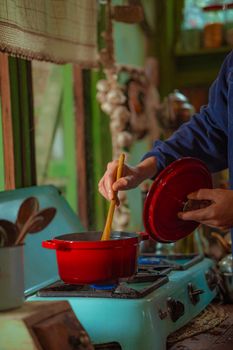 A person stirs food in a cozy kitchen using a red pot on a vintage stove.
