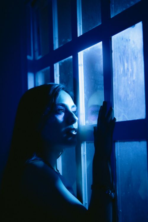 Free A moody portrait of a woman in blue light gazing out a window with a contemplative expression. Stock Photo
