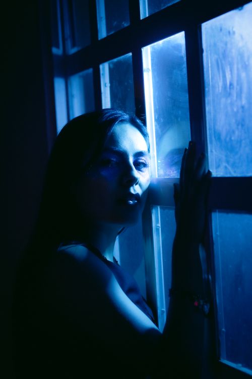 Free A dramatic portrait of a woman by a window with blue lighting. Stock Photo