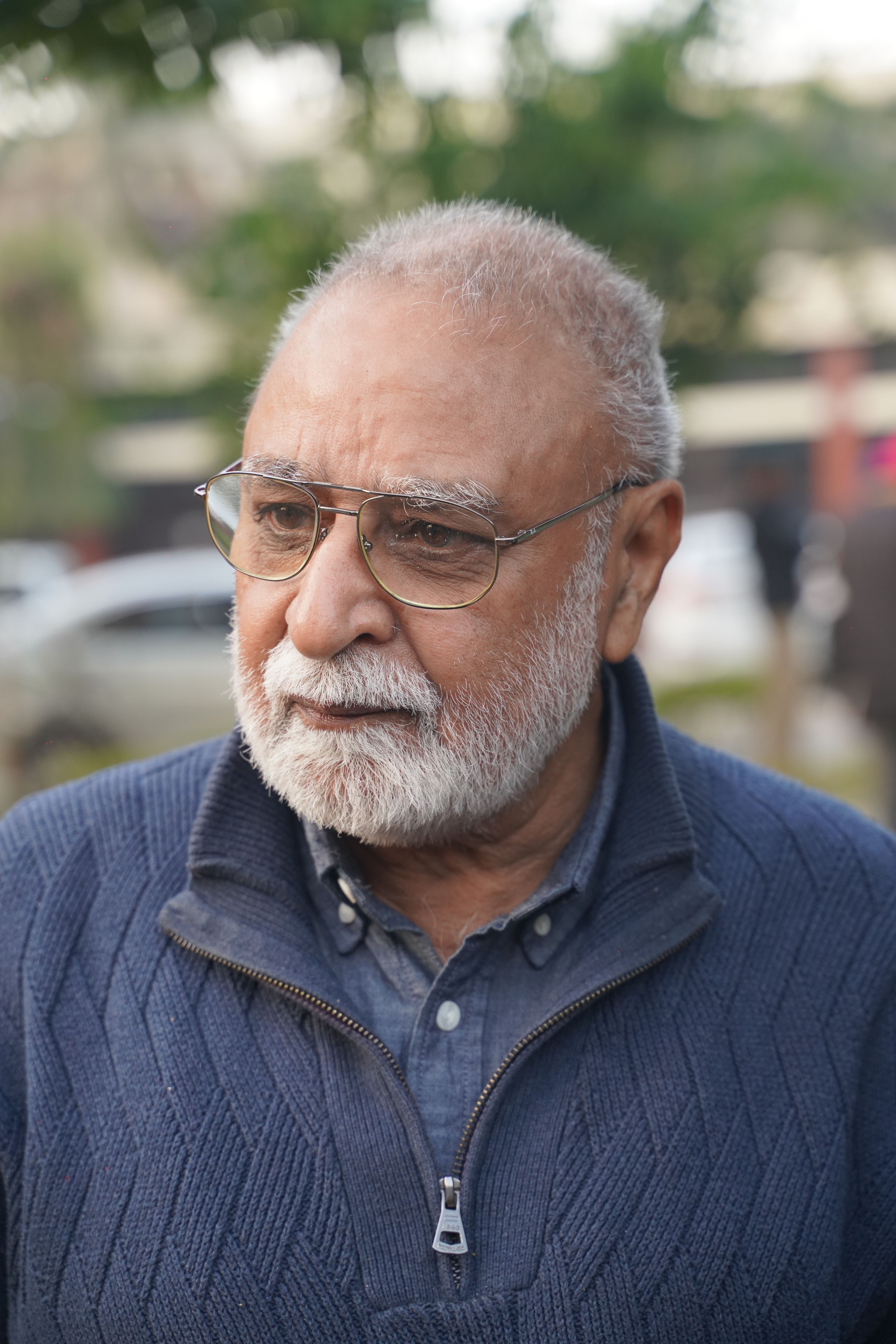 Close-up of an elderly man wearing glasses and a blue sweater in an outdoor setting.
