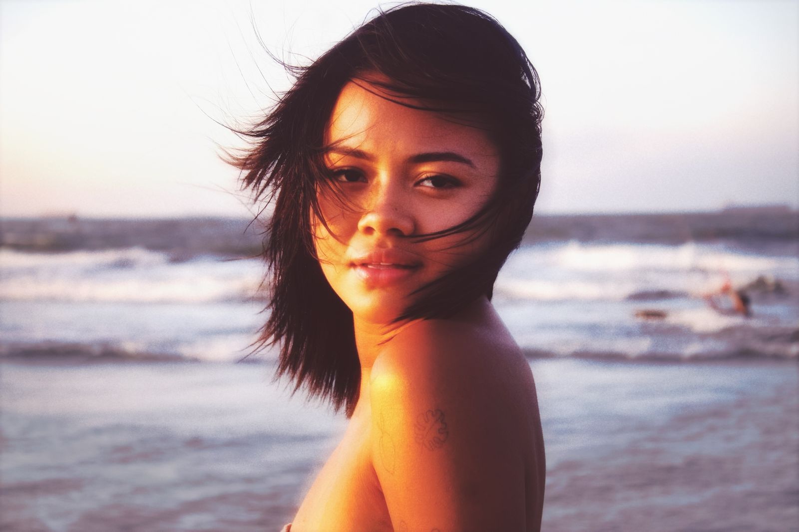Woman with windblown dark hair looking over her bare shoulder at a beach during golden sunset