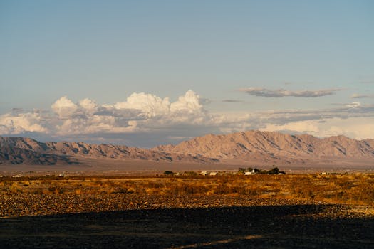 Broad desert vista under a vast sky, featuring distant mountain ranges and scattered clouds.