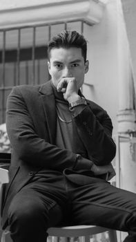 Confident young man in a black suit, seated indoors, exudes classic style in monochrome.
