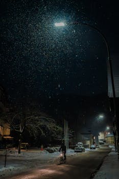 A tranquil night scene in Lilienfeld, Austria, featuring a cyclist on a snow-covered street under street lamps.