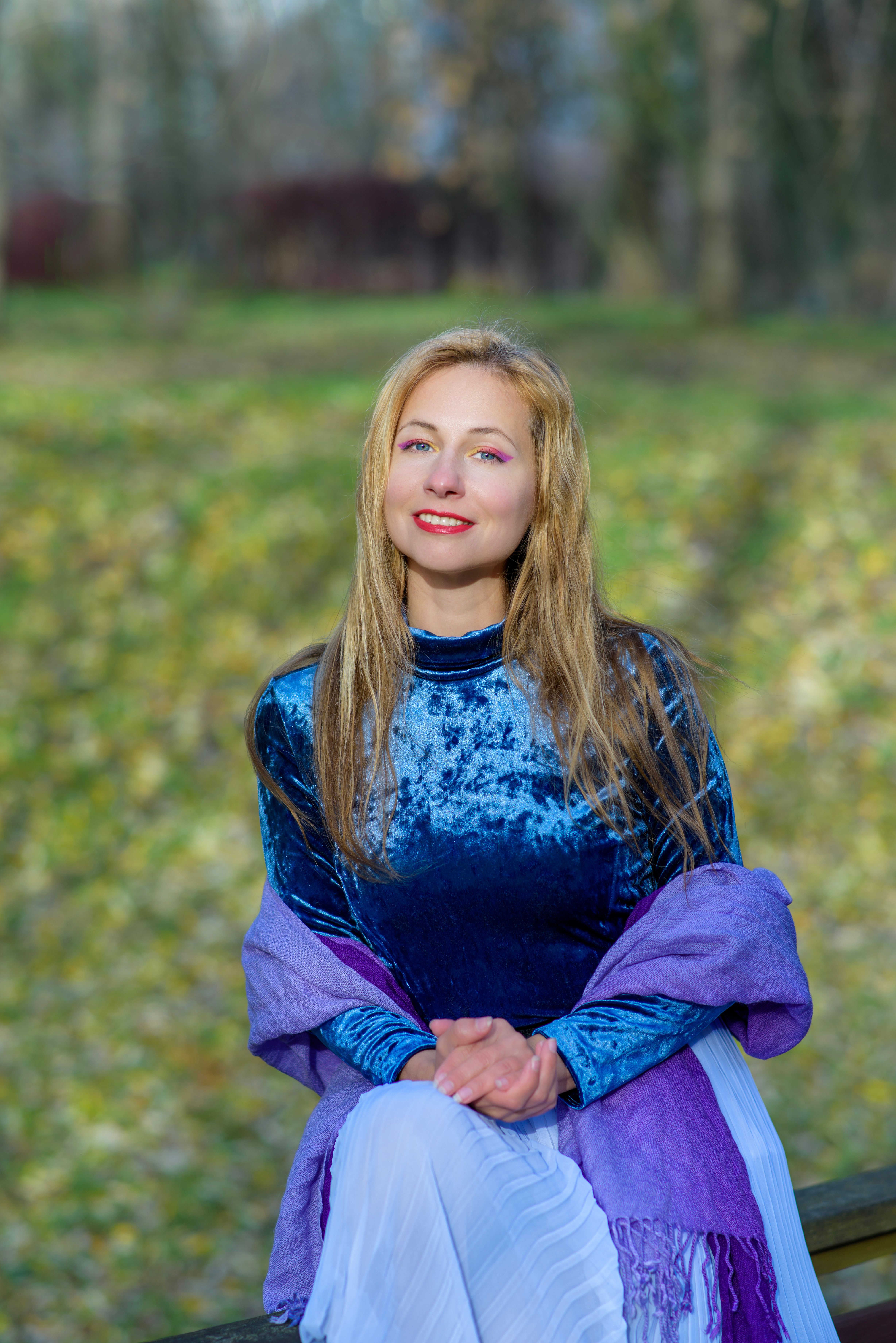 Elegant woman in blue velvet dress and purple scarf smiling outdoors in nature.