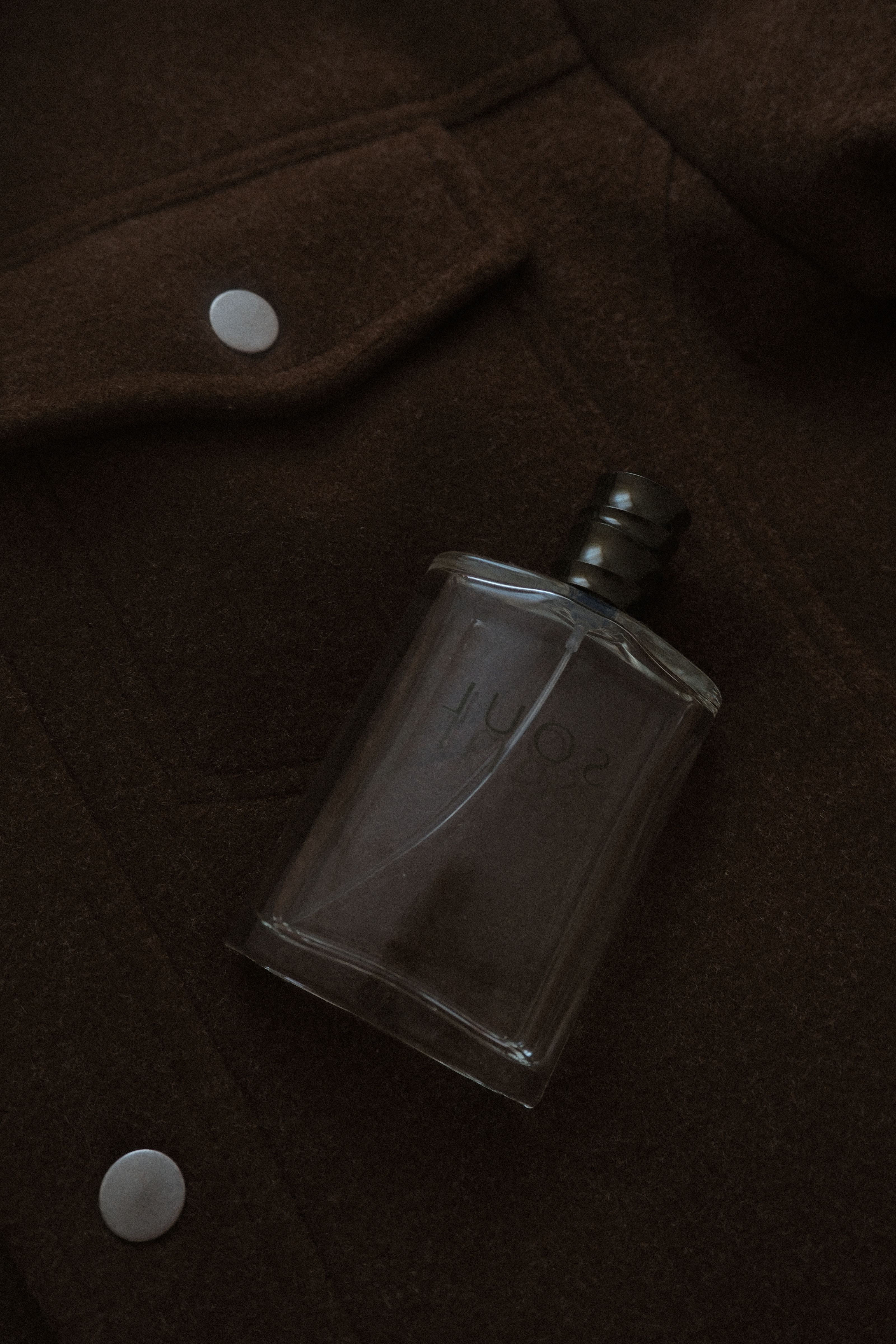 Frasco de perfume estiloso colocado sobre uma jaqueta de lã marrom escura, evocando sofisticação.