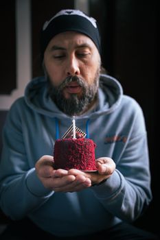 A man in a hoodie holds a small red velvet cake with a candle, celebrating a birthday indoors.
