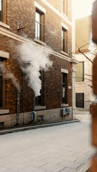 Steam rises from wall pipes in a city alley during daytime.