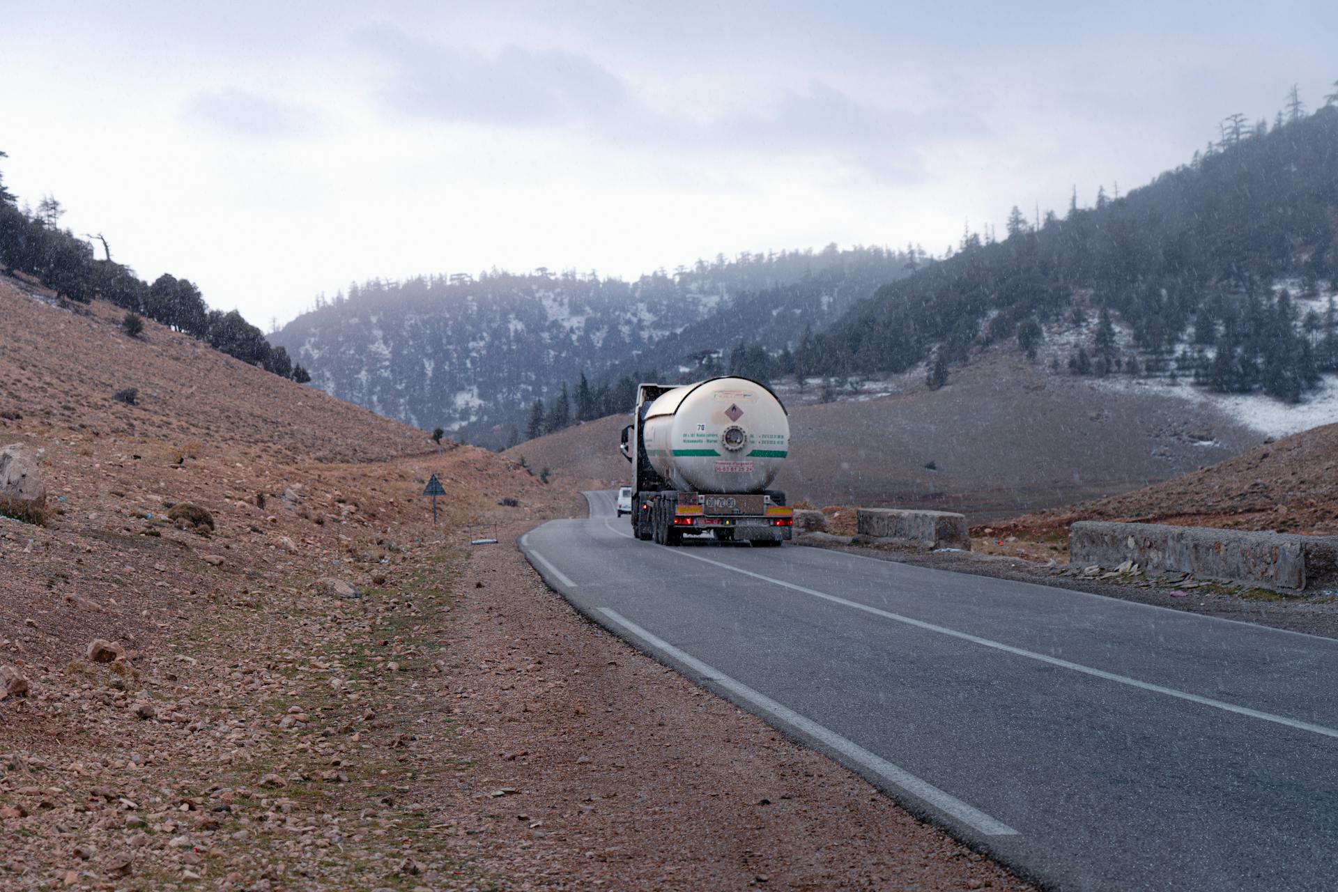 Gas tanker truck traveling on a mountainous road with snow-capped hills in the background.