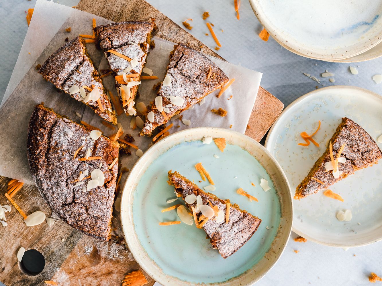 Sliced round orange-almond cake with a dark golden top, powdered sugar, sliced almonds and fine strips of orange zest on ceramic plates