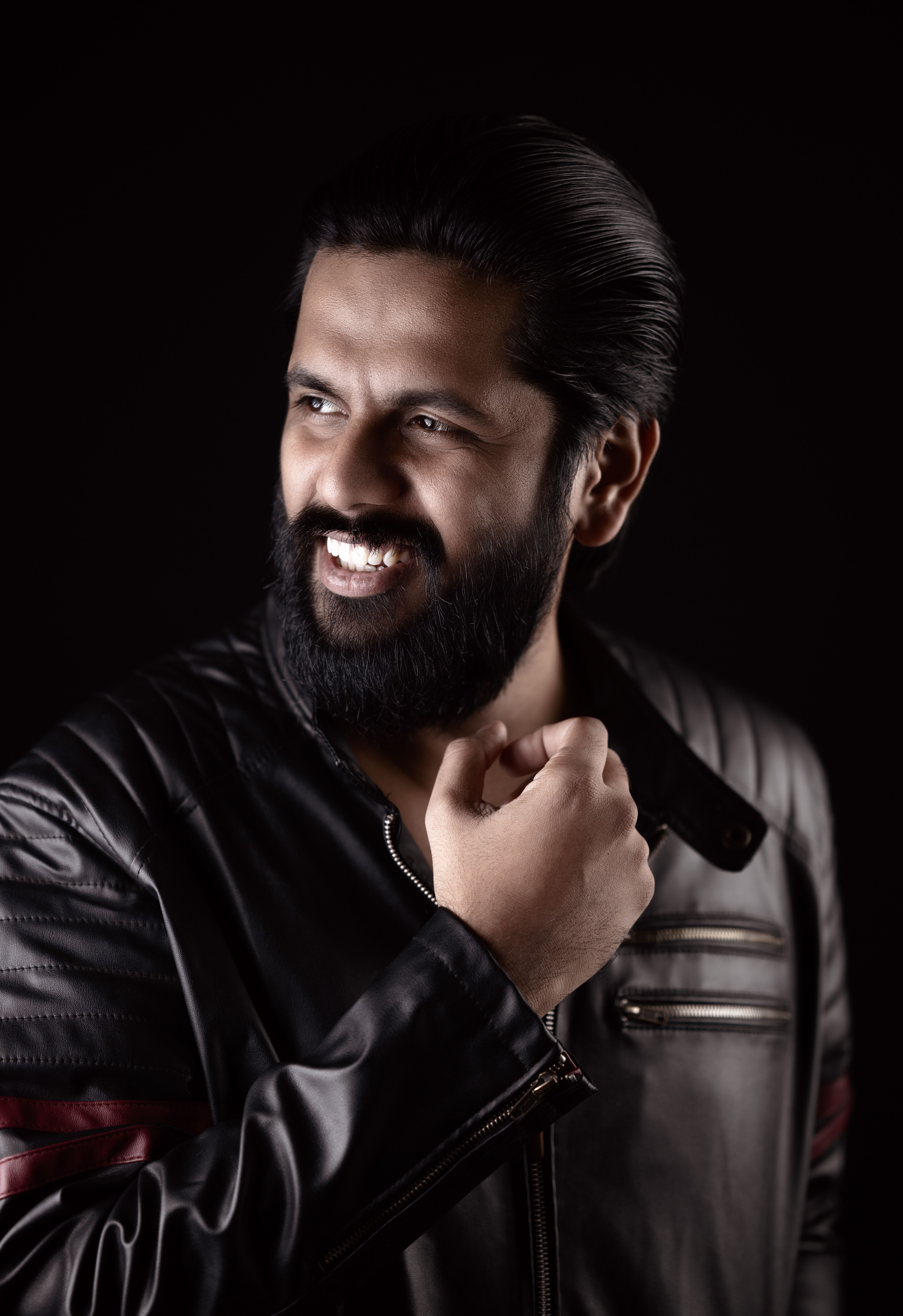 Portrait of a bearded man in a black leather jacket, smiling confidently against a dark background.