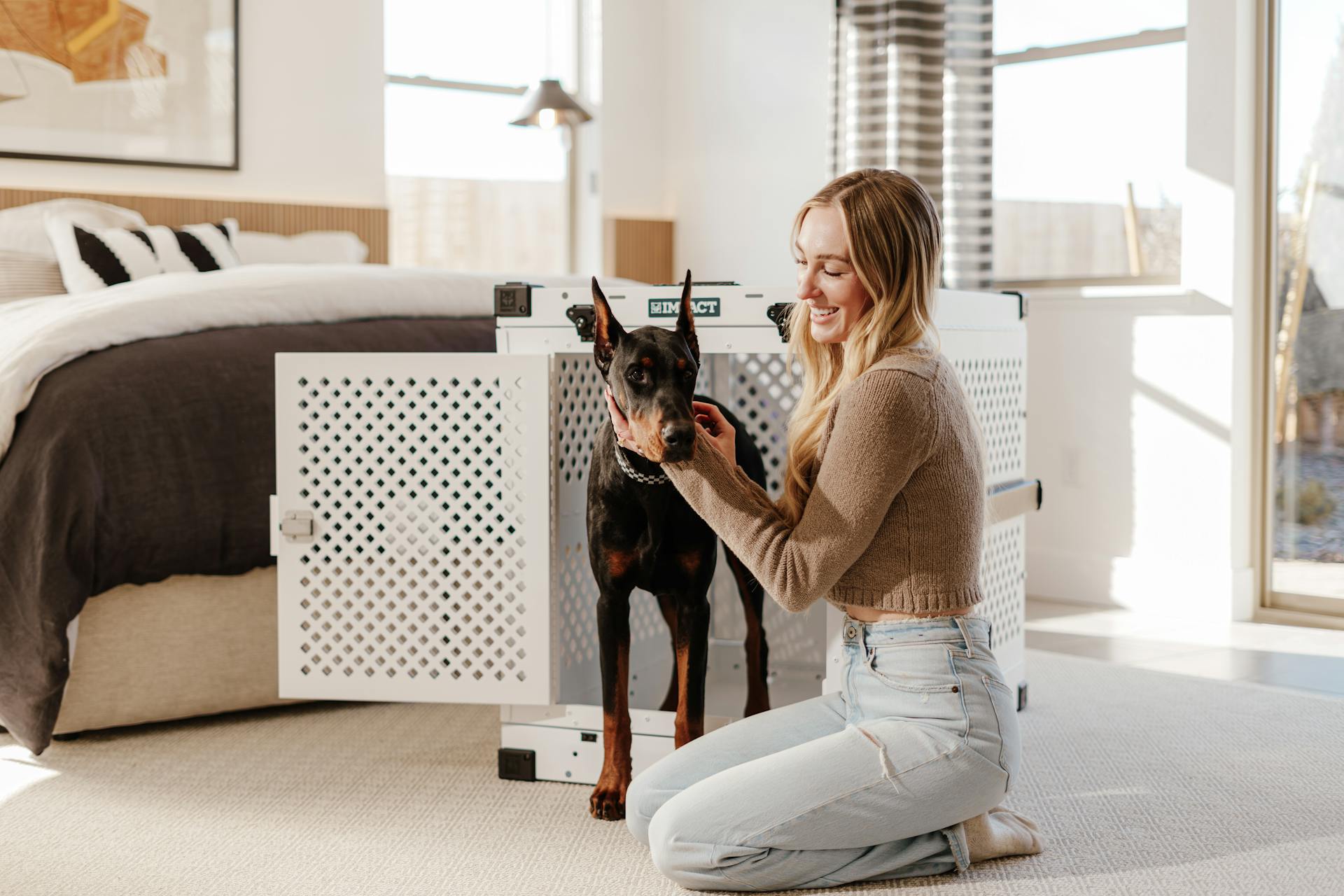 Young woman kneeling by Doberman and dog crate in a stylish bedroom setting.