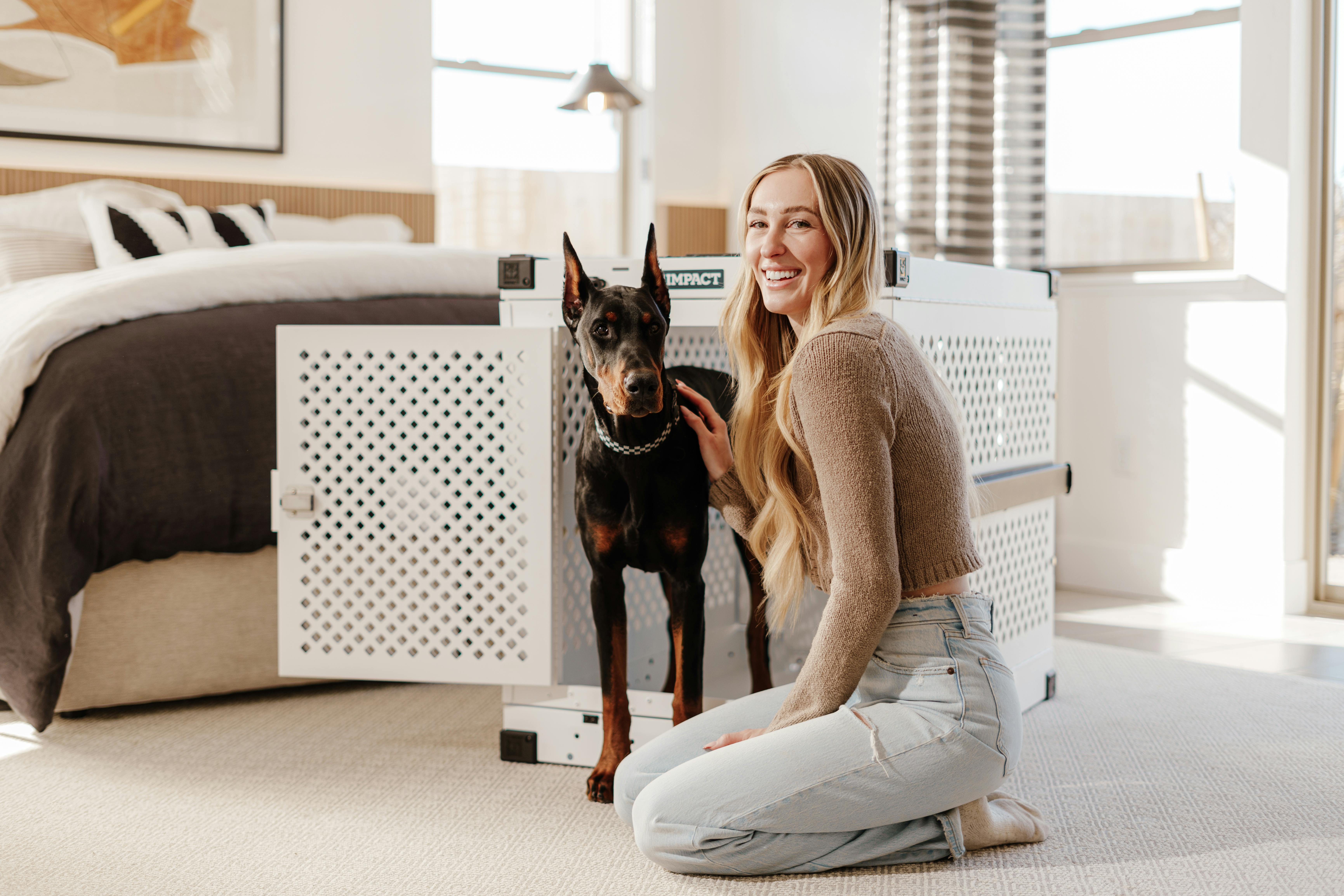A young woman smiling next to a Doberman inside a modern bedroom with natural light.