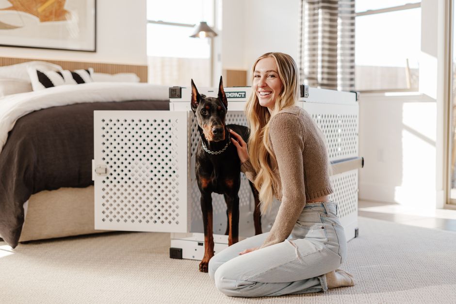 How Crate Training Solves Puppy House-Training Challenges A young woman smiling next to a Doberman inside a modern bedroom with natural light.