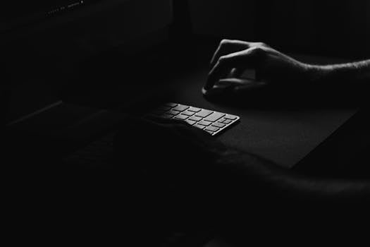 Moody black and white photo of hands typing on a keyboard in the dark, creating a mysterious atmosphere.