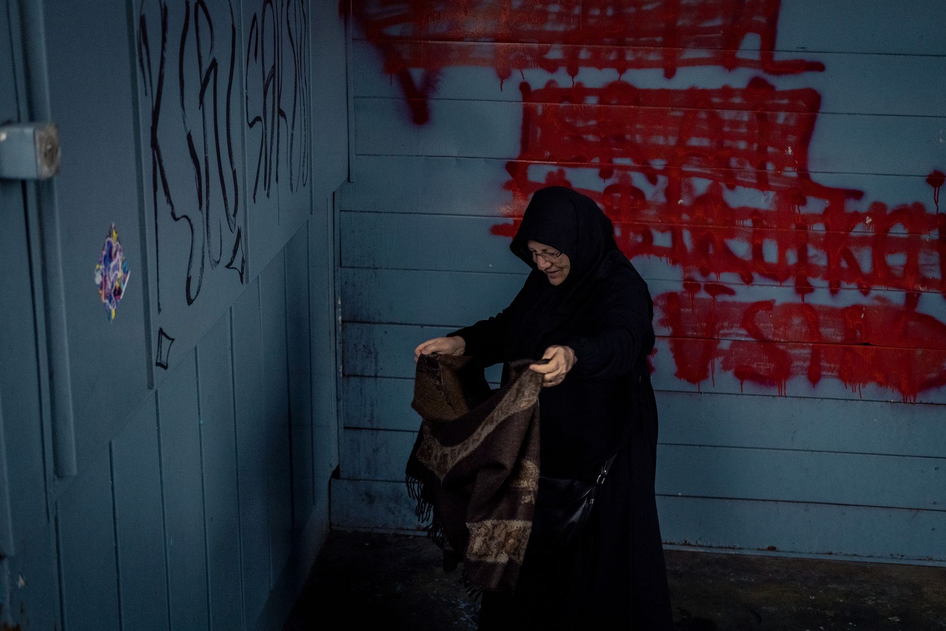 Elderly woman folding fabric against a graffiti-covered wall in an urban setting.