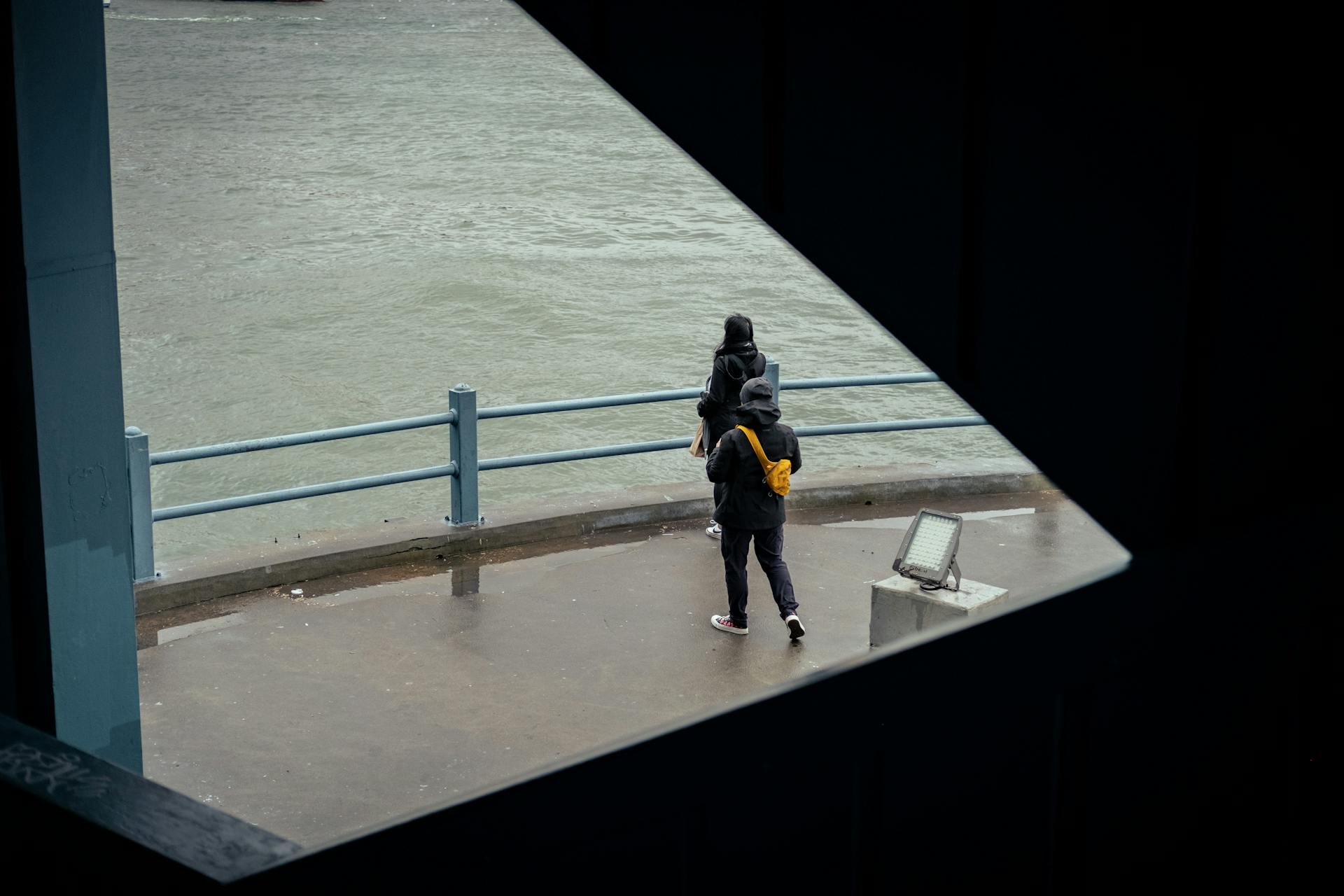 Two individuals stroll by a riverside on a cloudy day, viewed through an architectural frame.