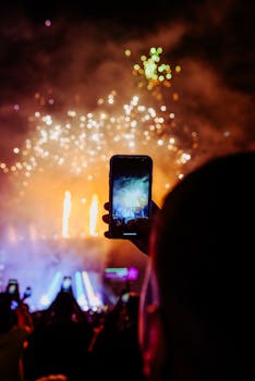 Vibrant fireworks illuminate the night sky, captured through a mobile camera at an outdoor celebration.