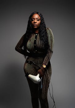 Elegant woman with long wavy hair holds a blow dryer in a studio setting, showcasing style and confidence.