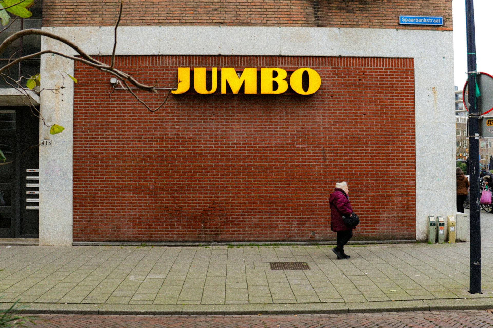 Elderly woman walking by Jumbo supermarket with red brick wall background in urban setting.