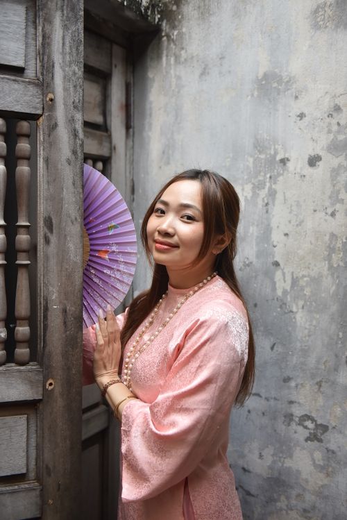 Free A young woman in a traditional pink dress holding a purple fan by a rustic doorway. Stock Photo