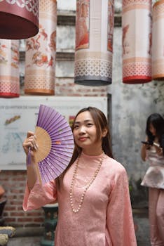 Elegant woman in traditional pink attire holding a fan, surrounded by lanterns.