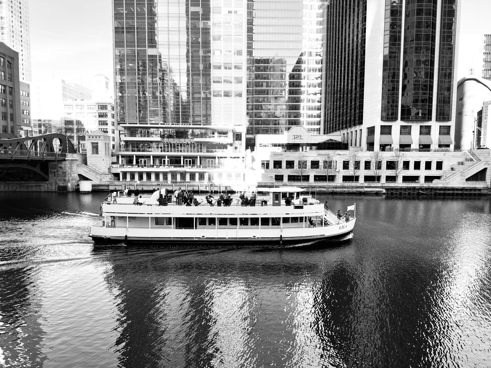 Black and white image of a riverboat cruising through an urban cityscape with skyscrapers.